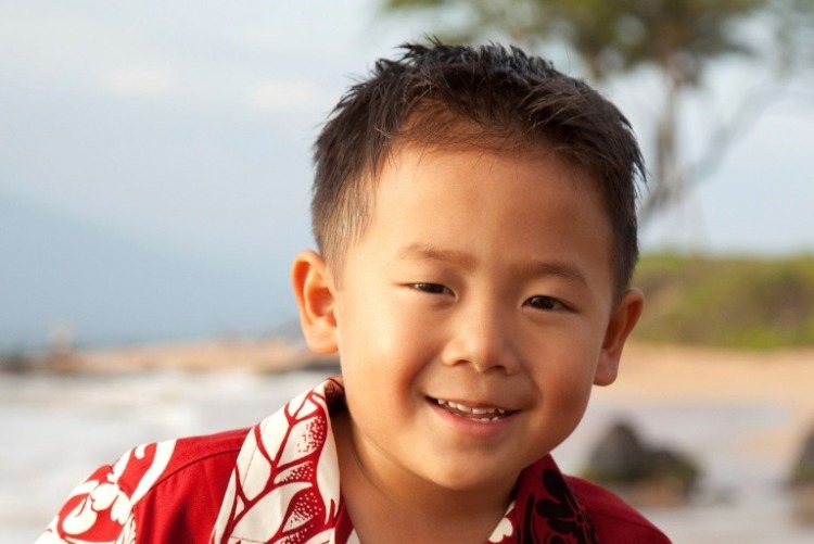 Smiling Asian boy with short brown colored hair, wearing a traditional button-up red and white Hawaiian shirt standing on a Hawaii beach.