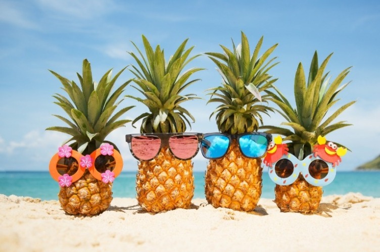 Four humorous looking pineapples decorated with tropical sunglass accessories, the pineapples are sitting alongside each other on a Hawaii beach.
