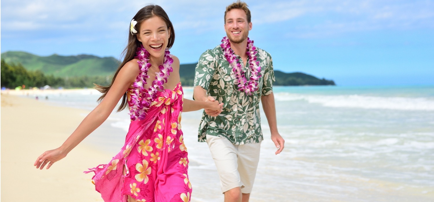 Handsome young man wearing khaki shorts and a green floral Hawaiian shirt. He is smiling at a pretty women wearing a pink tropical dress with yellow plumeria flowers. The couple is holding hands while strolling on a white sand beach in Hawaii.