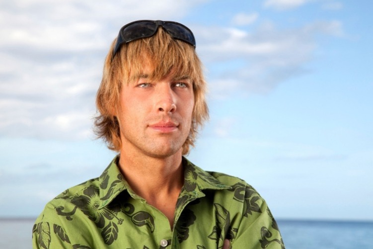 Good-looking young man standing on a beach with his arms folded, wearing sunglasses on his head, with the blue Pacific ocean in the background.. The man is donning a traditional green Aloha shirt.